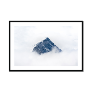 The image features a majestic, snow-capped mountain peak emerging from a dense layer of clouds against a pale sky, framed and ready for display.